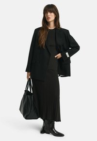 Schwarzer maßgeschneiderter Blazer mit doppelten Knöpfen, geripptem schwarzen Kleid, schwarzer Plissee-Rock und strukturiertem schwarzen Bag. Vervollständigt mit schwarzen Knöchelstiefeln.