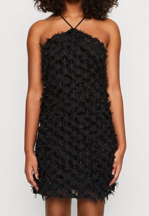 Vrouw draagt een mouwloze zwarte halterjurk met textuurrijke bloemapplicaties en subtiele pailletten, staand tegen een effen achtergrond.