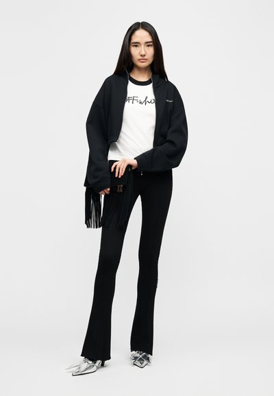 Μαύρο κοντό φούτερ με κουκούλα, λευκό μπλουζάκι με γραφικό σχέδιο, μαύρο καμπάνα παντελόνι και ασημένιες μυτερές γόβες. Λεπτομέρεια με φούντες στην τσάντα, μινιμαλιστικός σχεδιασμός.