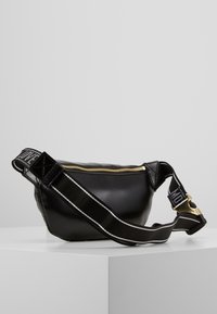 Schwarze Leder-Crossbody-Tasche mit einem Reißverschluss und einem strukturierten Tragegurt mit weißen Streifen. Goldene Beschläge an dem Reißverschluss.