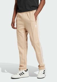 Beige track pants with white stripes, elastic waistband, side pockets, and embroidered logo. Paired with white sneakers featuring black stripes.