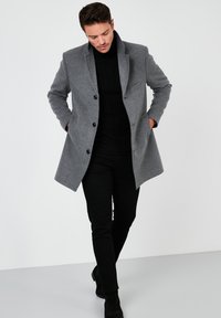 Abrigo de mezcla de lana gris con solapas con muescas, botones delanteros y bolsillos laterales. Llevado sobre un suéter de punto negro y combinado con pantalones negros y botas.