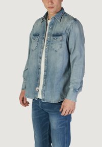Camicia di jeans azzurro chiaro con maniche lunghe, bottoni a pressione, due tasche sul petto e una texture leggermente sbiadita. Indossata sopra una maglietta bianca.