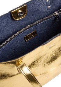 Sac fourre-tout doré avec intérieur en tissu bleu marine, poche zippée, fermeture à pression et "W.11" embossé sur les étiquettes métalliques à l'intérieur et à l'extérieur.