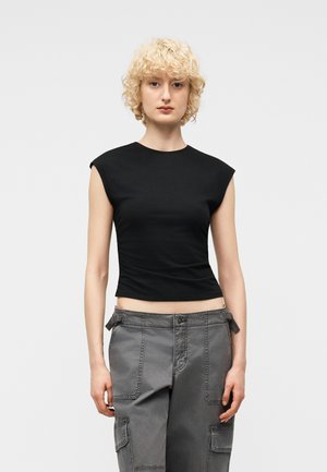 Persoon met kort, krullend blond haar, gekleed in een zwart mouwloos topje en grijze cargobroek, staand tegen een effen lichte achtergrond.