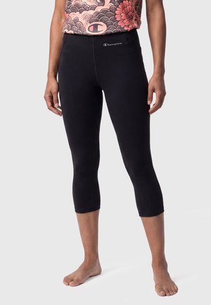 Frau trägt schwarze kurze Leggings mit Champion-Logo nahe der Taille und ein gemustertes Oberteil, steht barfuß vor einem einfarbigen Hintergrund.