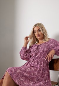 Purppura paisley-mekko, jossa on pitkät hihat, kerätty vyötärö ja valuva hame. Silkkinen kangas, istuu puisella tuolilla.