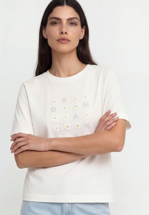 Frau mit langen dunklen Haaren, die ein weißes T-Shirt mit Gänseblümchen und Buchstaben trägt, die das Wort "ROMAN" über der Brust bilden.