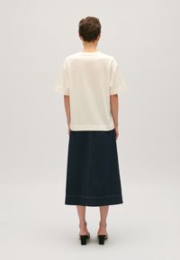 Henkilö seisoo selkä kameraan, yllään valkoinen lyhythihainen paita, tummansinisestä denimmistä valmistettu midi-hame ja mustat korkokenkä-sandaalit yksinkertaisella taustalla.