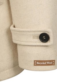 Beige gerecycled wollen jasje manchette met een gestructureerde afwerking, voorzien van een zwarte knoop en een label met de tekst "Gerecycled Wol" in witte letters.