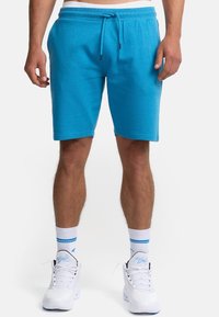 Shorts turquoise en tissu doux qui évacue la transpiration. Caractérisés par une taille élastique avec cordon de serrage. Texture lisse et coupe au niveau des genoux.