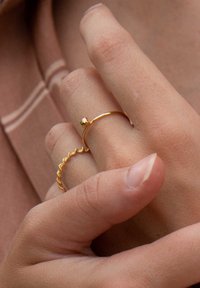 Goldene Ringe an einer Hand; einer ist ein glatter Bandring mit einer kleinen Kugel, der andere hat ein wellenförmiges, strukturiertes Design. Der Hautton ist hell.