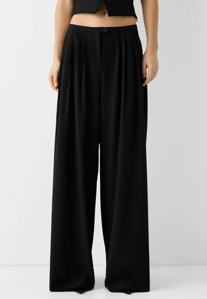 Pantalon noir à jambes larges avec plis à l'avant, fermeture par bouton et zip, porté avec un haut court noir, visible de la taille vers le bas.
