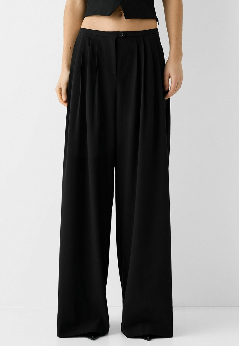 Pantalon noir à jambes larges avec plis à l'avant, fermeture par bouton et zip, porté avec un haut court noir, visible de la taille vers le bas.