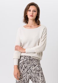 Crème brei trui met een bootnek en geribbelde textuur, voorzien van knopen op de manchetten; gecombineerd met beige broek in een zebrapatroon.