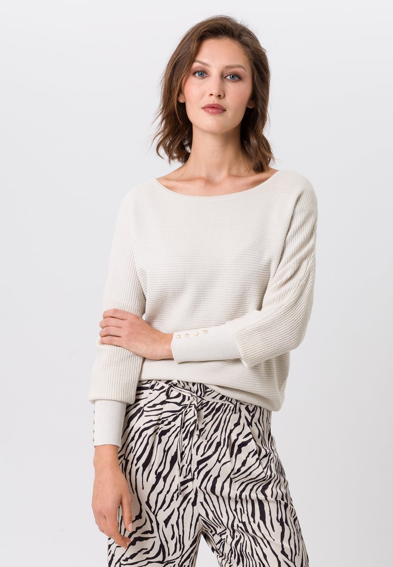 Crème brei trui met een bootnek en geribbelde textuur, voorzien van knopen op de manchetten; gecombineerd met beige broek in een zebrapatroon.