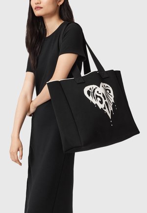 Sort tote taske lavet af robust stof, med et hvidt hjerte og abstrakt design, to håndtag og en rummelig rektangulær form.