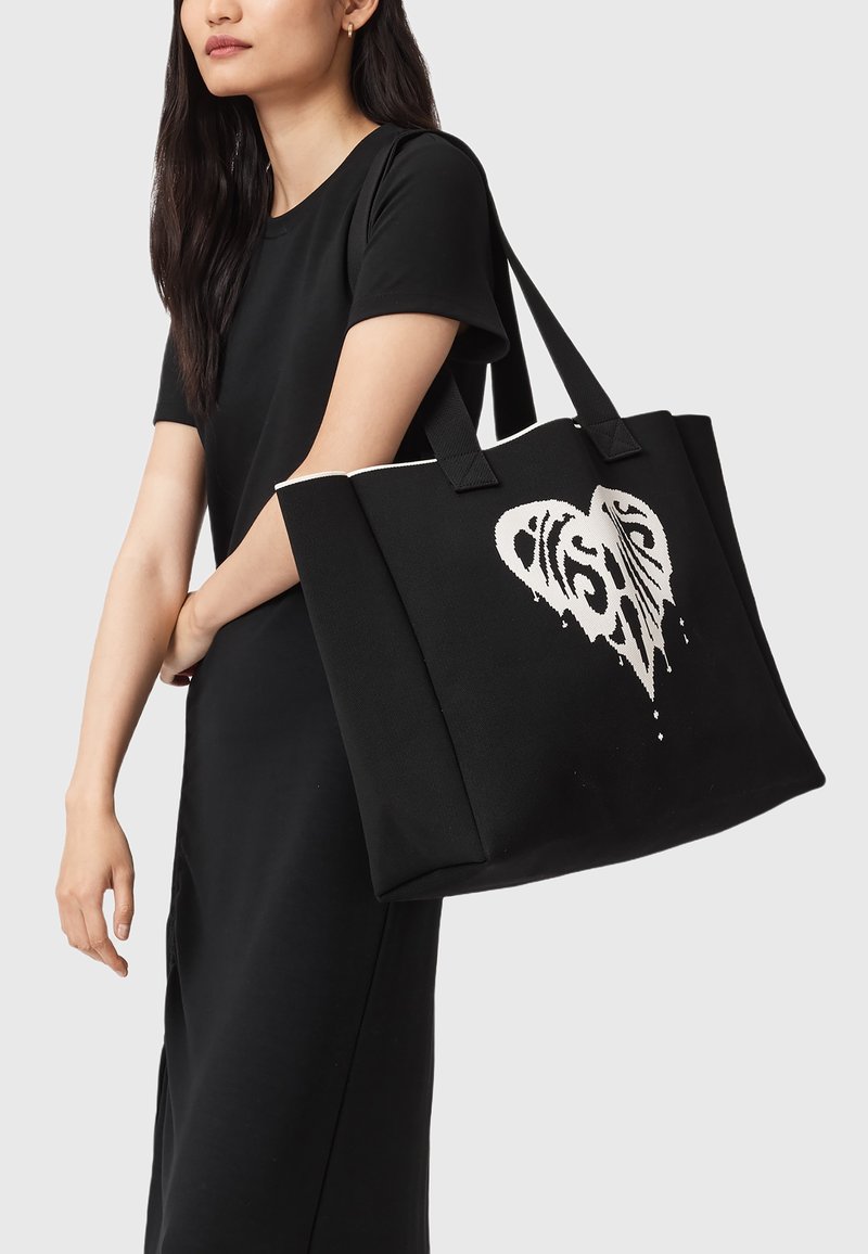 Sort tote taske lavet af robust stof, med et hvidt hjerte og abstrakt design, to håndtag og en rummelig rektangulær form.