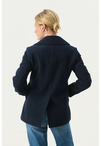 Marinblå ullkappa med bred krage, enkel slits bak och skräddarsydd passform, i kombination med ljusblå jeans.