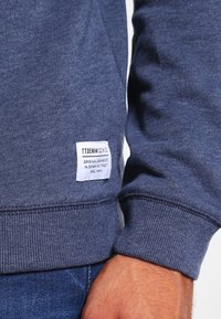 Marinblå sweatshirt gjord av mjukt, texturerat tyg. Har ribbade ärmslut och fåll, med en rektangulär vit etikett som anger varumärket.
