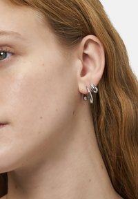 Silberne, metallische Ohrringe mit einer flüssigen, abstrakten Form. Sie verfügen über eine glänzende Oberfläche und zwei deutlich miteinander verbundene Elemente.