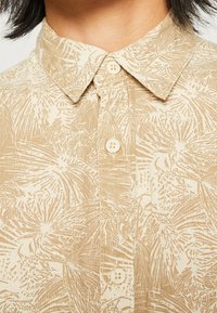 Chemise à manches courtes beige avec un imprimé botanique. Dotée d'un col à boutons et de boutons assortis. Tissu texturé avec un aspect naturel.