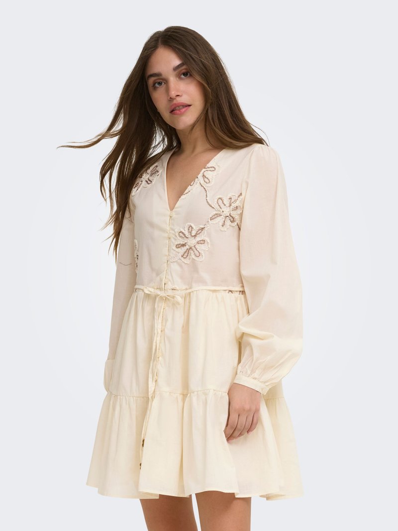Femme aux longs cheveux bruns portant une robe crème à manches longues avec broderies florales et une jupe à volants, debout devant un arrière-plan uni.