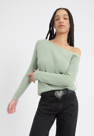 Lichtgroene geribde gebreide trui met off-shoulder ontwerp, lange mouwen en een zwarte riem met een hartvormige gesp. Draagt het met zwarte jeans.