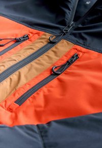 Jachetă colorată în blocuri, având panouri portocalii, maro și negre, cu trei buzunare cu fermoar și țesătură netedă, texturată.