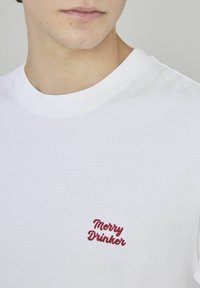 T-shirt en coton blanc avec col rond, portant un texte brodé rouge "Merry Drinker" sur la partie gauche de la poitrine. Texture douce.