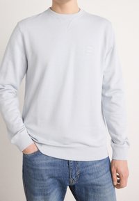 Homme portant un sweat-shirt gris clair uni à col rond et un jean bleu, une main dans la poche, sur un fond uni.