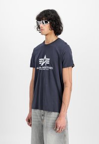 Merimi t-paita tummansinisena, jossa on valkoinen logo, lyhyet hihat ja rento istuvuus, yhdistettynä vaaleanharmaisiin farkkuihin ja urheilullisiin valkoisiin aurinkolaseihin.