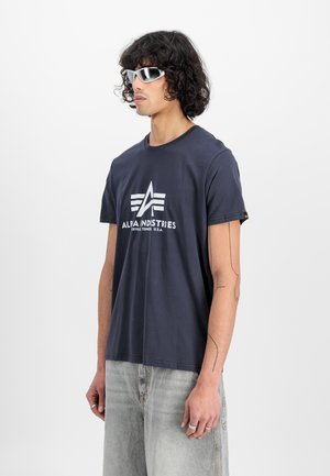 Navy cotton t-shirt with a white logo, short sleeves, and a relaxed fit, paired with light grey denim jeans and sporty white sunglasses.