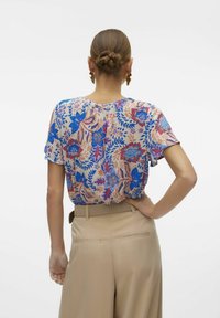 Bluse mit kurzen Ärmeln im Blumenmuster, die blaue und rote Muster auf beigem Hintergrund zeigt. Kombiniert mit beigen Hosen.