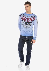 Blaues langärmliges T-Shirt mit dem grafischen Design "LEGEND 1996", getragen mit dunklen Jeans und weißen Sneakers.