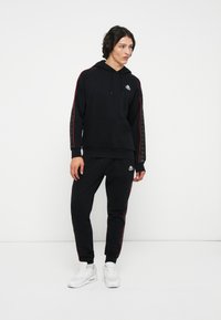 Joven vestido con sudadera negra y pantalones deportivos con rayas rojas y zapatillas blancas, de pie frente a un fondo blanco.