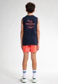 Granatowy top bez rękawów z czerwonym napisem; jasne koralowe szorty z małymi granatowymi wzorami kotwic; białe trampki z zielonymi akcentami.