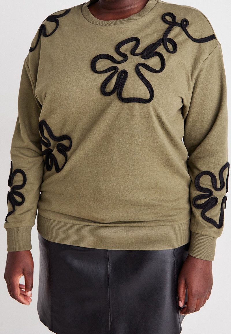 Sweat-shirt vert olive avec des applications florales abstraites noires sur la poitrine et les manches, porté avec une jupe en cuir noire.