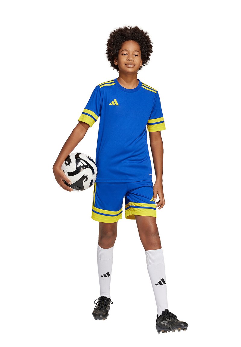 Blaues Fußballtrikot mit einem blauen Shirt mit gelben Akzenten, passenden Shorts, weißen Socken und schwarzen Fußballschuhen. Der Spieler hält einen schwarz-weißen Fußball.