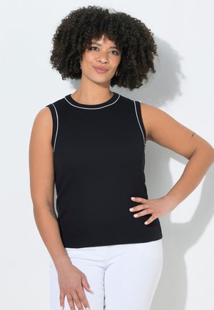 Femme aux cheveux bouclés portant un haut noir sans manches et un pantalon blanc, debout avec une main sur la hanche devant un fond blanc uni.