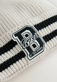 Beige gestrickte Mütze mit schwarzen Streifen und einem auffälligen schwarzen und weißen gestickten Patch mit einem "B" und dem Text "Baker".