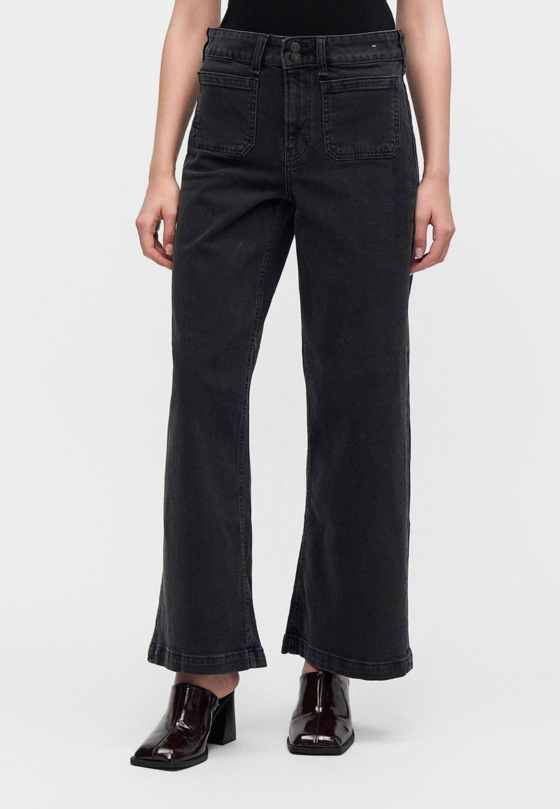 Personne portant un jean noir taille haute à jambes larges avec poches plaquées devant et chaussures marron foncé à talons carrés sur un fond uni.