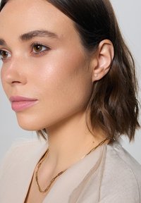 Złote kolczyki w kształcie kwiatów, gładka faktura, w zestawie ze złotym naszyjnikiem w kształcie spinacza. Odcień skóry jest neutralny; włosy są ciemnobrązowe i falowane.