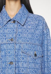 Primer plano de una persona que lleva una chaqueta de mezclilla azul con un patrón repetido de texto "MOSCHINO" y botones negros.
