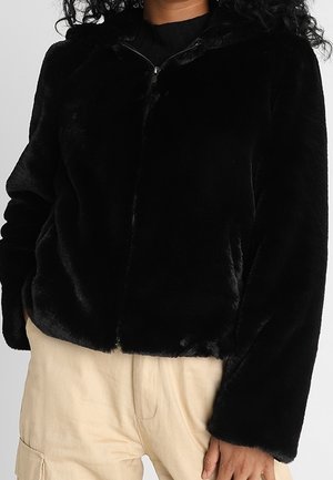 Vrouw draagt een zwart, pluizig vest met rits en beige broek, met hand in de zak, gefotografeerd van nek tot heup tegen een effen achtergrond.