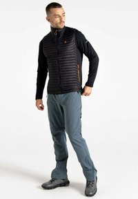 Schwarze Daunenweste mit gestepptem Design, Reißverschlusstaschen und orangefarbenen Akzenten. Kombiniert mit hellgrauen Hosen und robusten schwarzen Stiefeln.