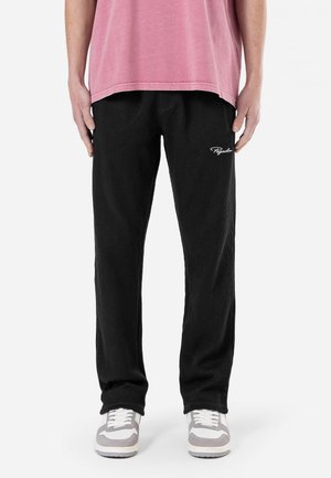 Hombre con pantalones de pana negros con pequeño logo blanco en el muslo, camisa rosa y zapatillas blancas, de pie frente a un fondo liso.