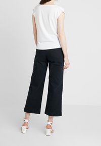 Camiseta blanca ajustada de manga corta, combinada con pantalones anchos negros y sandalias blancas de tacón cuadrado, vista desde atrás.