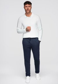 Homme debout portant un polo blanc à manches longues, un pantalon de survêtement bleu marine à cordon de serrage et des baskets blanches, contre un fond clair et uni.