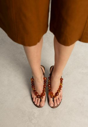 Voeten met bruine sandalen versierd met grote barnstenen kralen, staand op een lichtgrijze vloer, met een bruine rok zichtbaar boven de knieën.